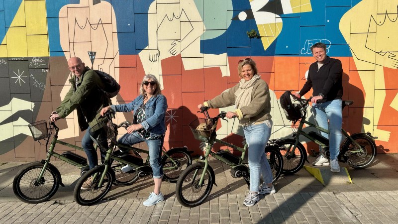 Four people on bikes pose by a colorful mural on a sunny day.