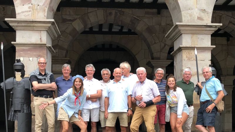 Group of people posing in front of a stone archway with spears displayed nearby.