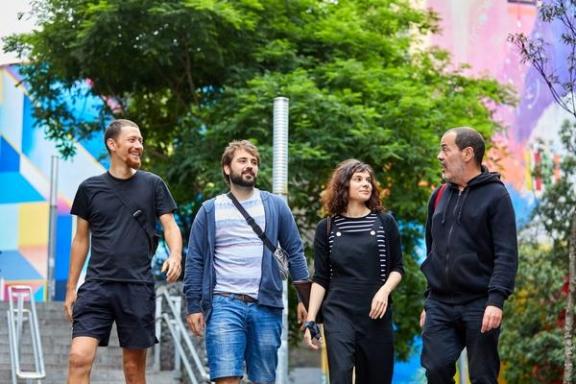 Four people smiling and walking outdoors, with colorful murals and trees in the background.