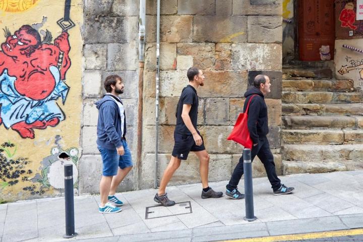Three people walking on a sidewalk past mural artwork on a wall.