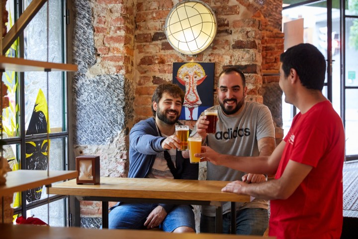 Three men clinking glasses of beer at a wooden bar table in a brick-walled pub.