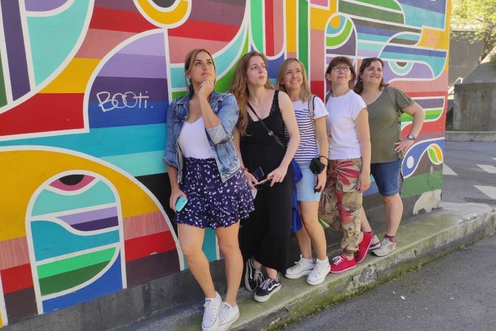 Five people pose playfully in front of a colorful, geometric mural on a street.