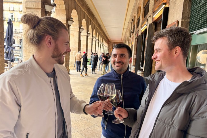 Three men smiling and toasting with red wine glasses under an archway.