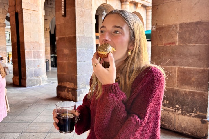 Woman in a red sweater eats pastry and holds a drink under stone arches.