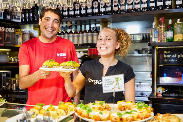 Two smiling people holding plates of food in a bar setting.