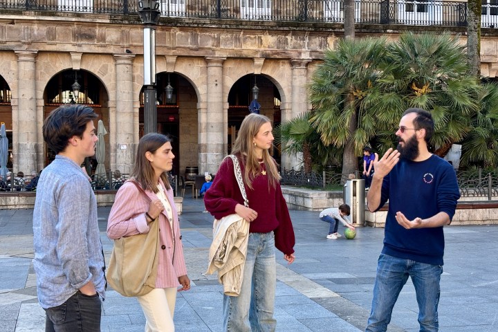Four people talking in a plaza with arches and palm trees in the background.