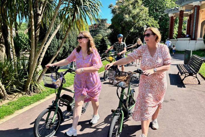 Two women in floral dresses walk bicycles on a sunny park path.