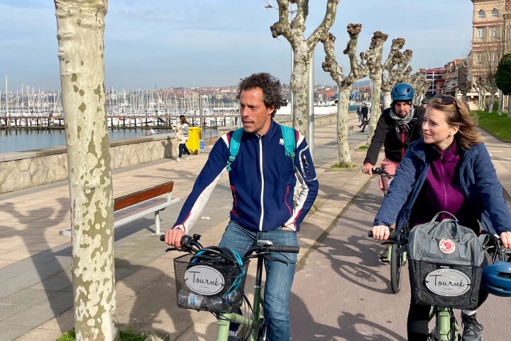 People cycling on a path lined with pruned trees near a marina on a sunny day.