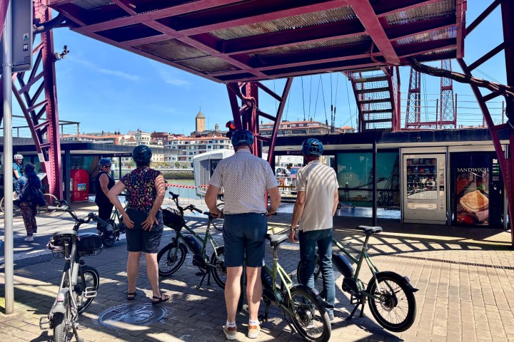 People with bikes stand under a red bridge with cityscape in the background.