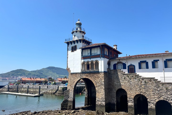 Historic lighthouse building with stone arches by the water under a clear blue sky.