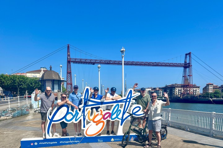 Group posing with 'Portugalete' sign near a river and suspension bridge under clear blue sky.