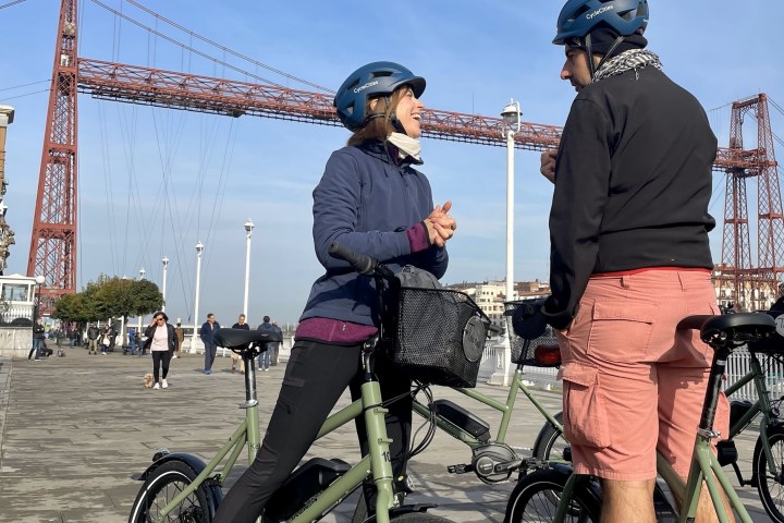 Two cyclists talking near a red iron bridge on a sunny day.