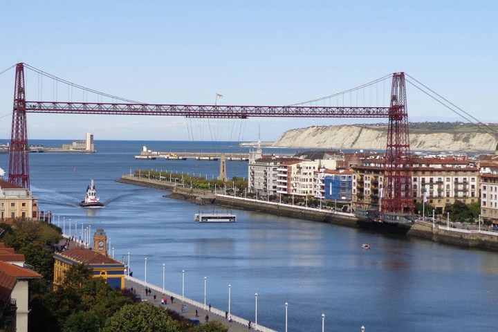 Suspension bridge over a river with cityscape and hills in background.