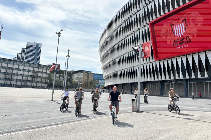 People riding bikes near a modern stadium with a large red sign.