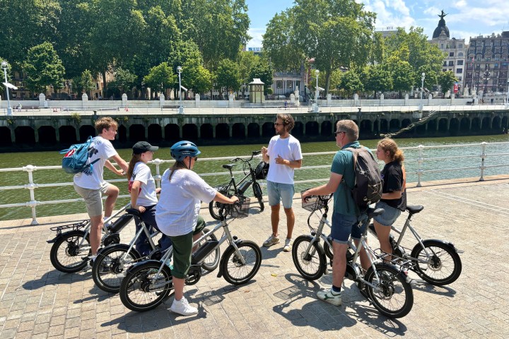 Group of people with bikes gathered on a riverside promenade under a sunny sky.