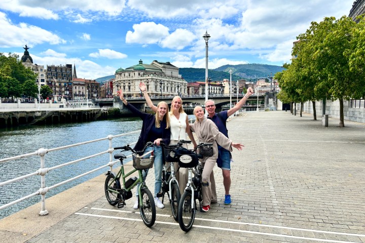 Four people with bicycles posing by a riverside promenade with buildings and mountains in the background.
