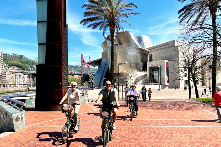Cyclists riding in front of a modern building with palm trees on a sunny day.