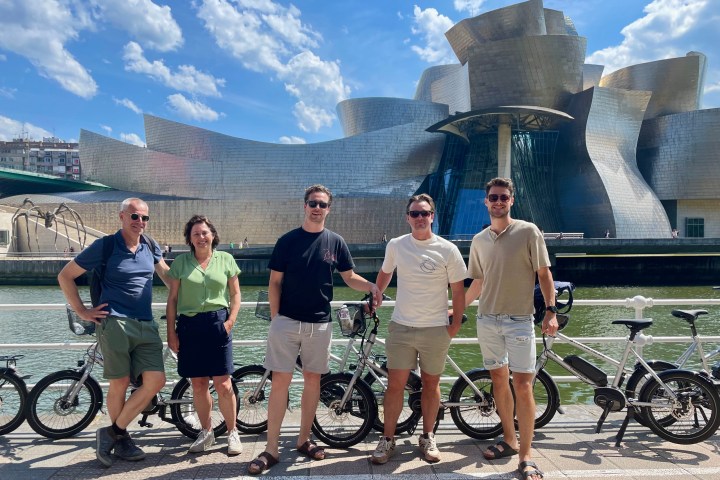 Five people with bikes in front of a modern, metallic building with cloudy sky.