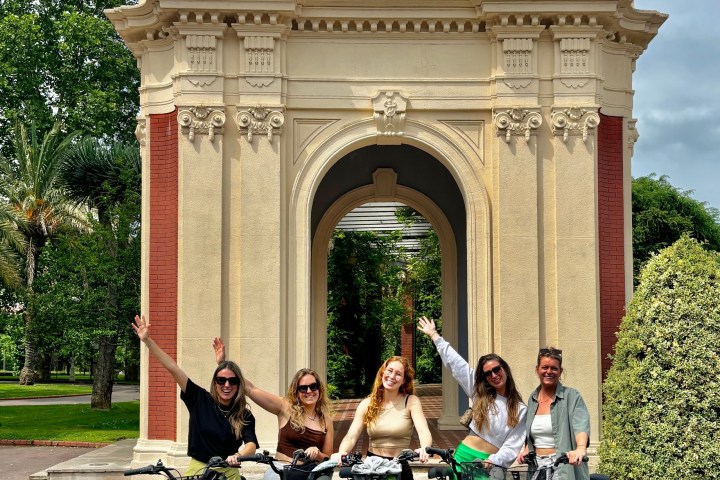 Five people on bikes posing in front of a decorative arch in a park.