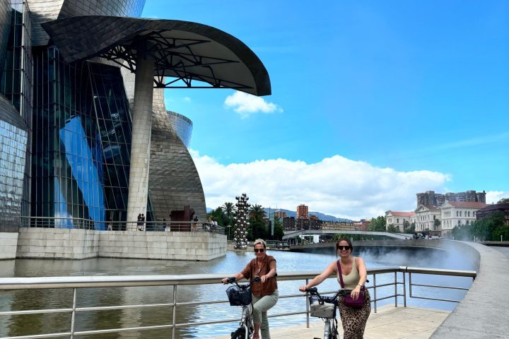 Two people biking near modern building by water under blue sky.
