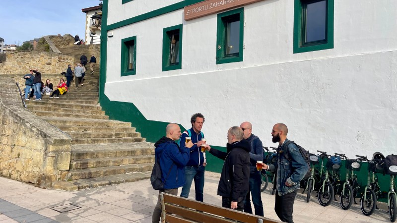 Group of men chatting near 'Portu Zaharra Taberna' with people sitting on nearby steps.