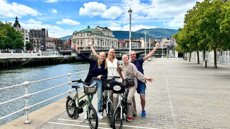 Four people with bicycles posing by a riverside with a historic building and mountains in the background.