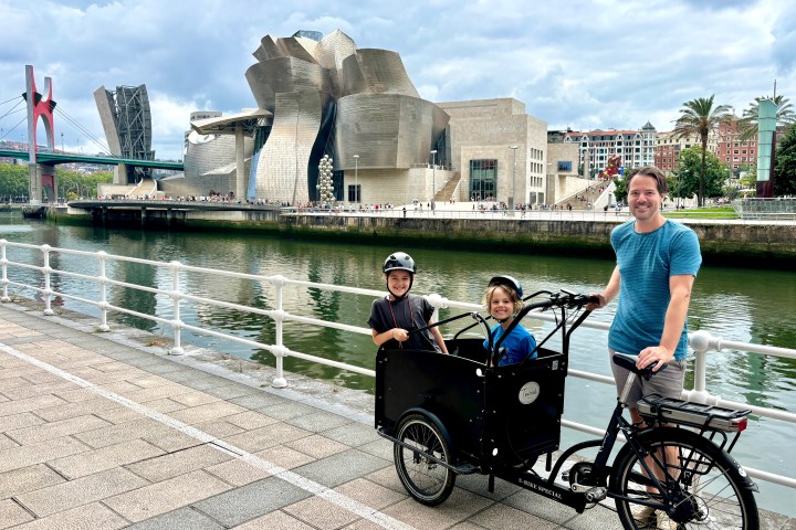 Person with two children in a cargo bike near a museum and river walkway.