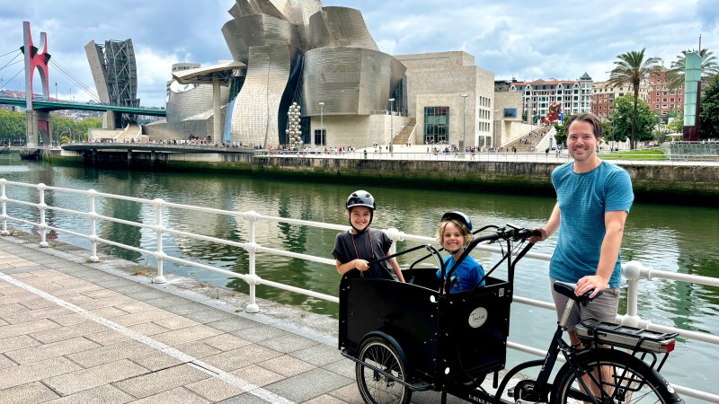Person with two children in a cargo bike near a museum and river walkway.