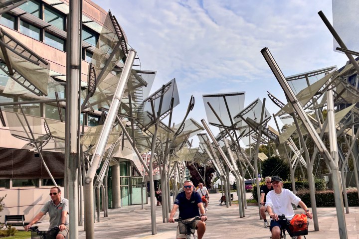 Three people cycling under metal sculptures in an urban area with a cloudy sky.