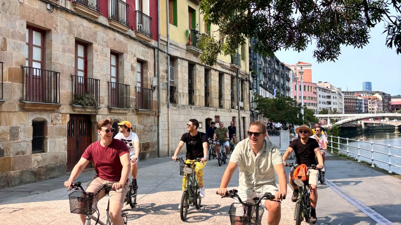 Group of cyclists riding on a riverside path next to colorful buildings.