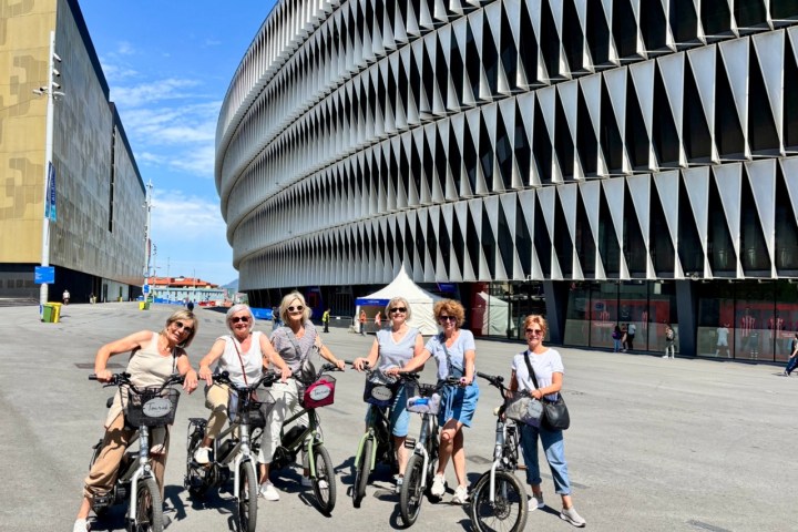 Six people on bicycles pose in front of a modern building with unique architecture.
