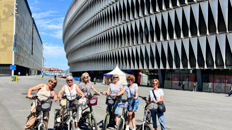 Six people on bicycles pose in front of a modern building with unique architecture.