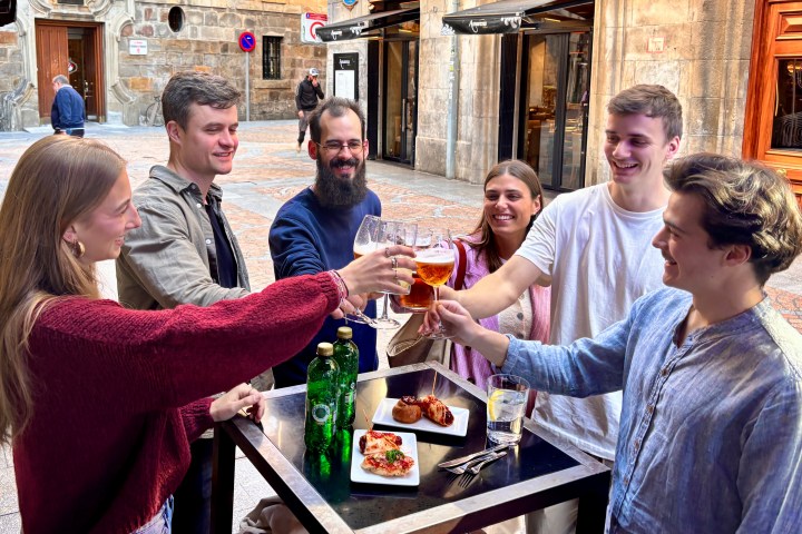 Six people cheer with drinks around a table on a street patio.