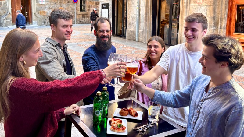 Six people cheer with drinks around a table on a street patio.