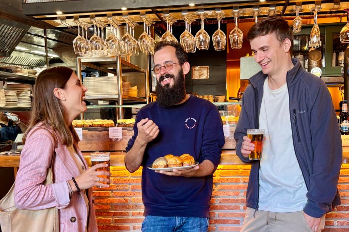 Three people in a bar, smiling and holding drinks and snacks.