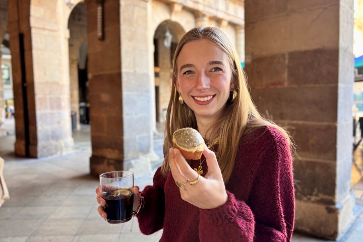 Woman smiling, holding a drink and a pastry in an outdoor setting.
