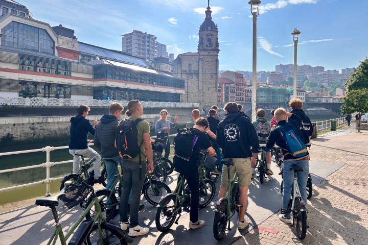 Group of people with bikes near a river, historic building in the background under a sunny sky.