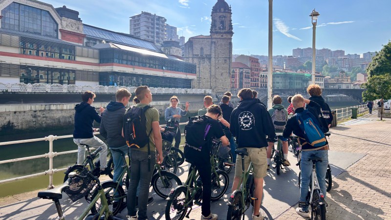 Group of people with bikes near a river, historic building in the background under a sunny sky.
