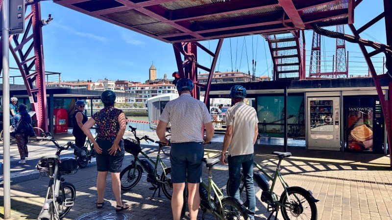 Cyclists with helmets and bikes under a red metal bridge, cityscape and vending machines in background.