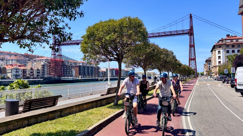 Five cyclists ride along a riverside bike path under a red suspension bridge on a sunny day.