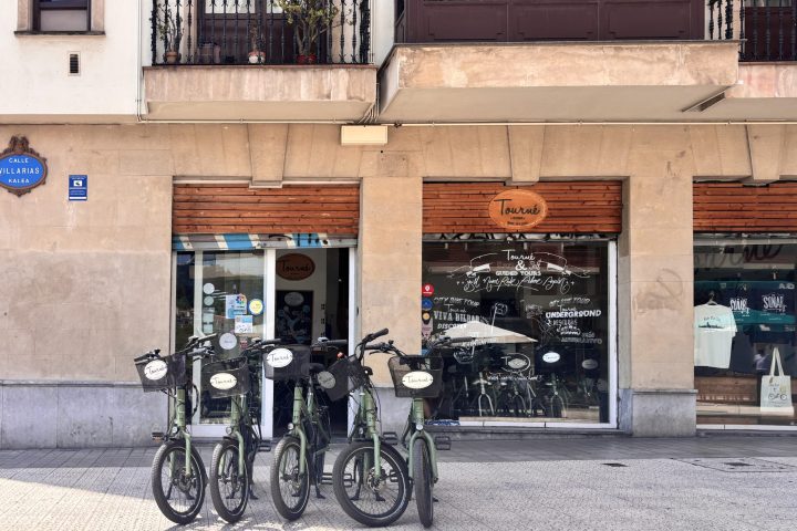 Four bicycles parked in front of a storefront with large windows and a balcony above.