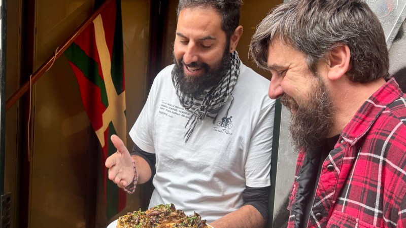 Two men smiling and holding a plate of food, Basque flag in the background.