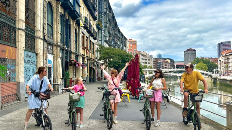 Five people on bicycles in a city street near a river, with colorful buildings and a cloudy sky.