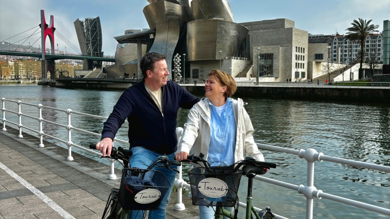 Two people with bikes by a riverside, Guggenheim Museum Bilbao in the background.