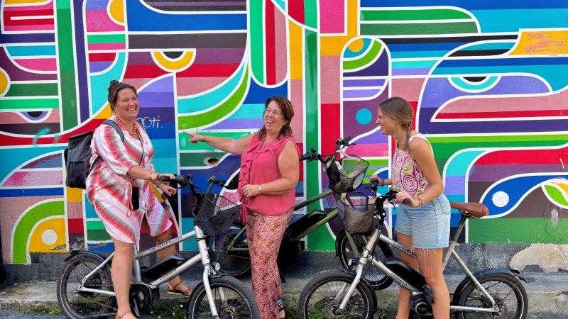 Three women with bikes smiling in front of a colorful mural.