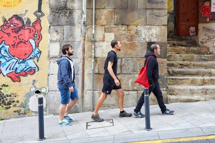 Three people walking by colorful street art on a stone wall.
