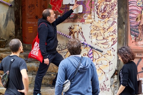 Guide with red bag explains graffiti to three tourists on steps.