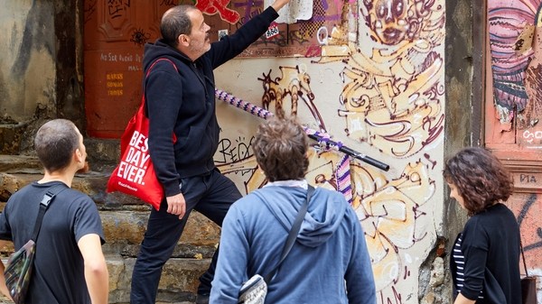 Guide with red bag explains graffiti to three tourists on steps.