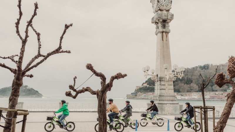 Five people riding bikes in front of a seafront clock tower on a cloudy day.