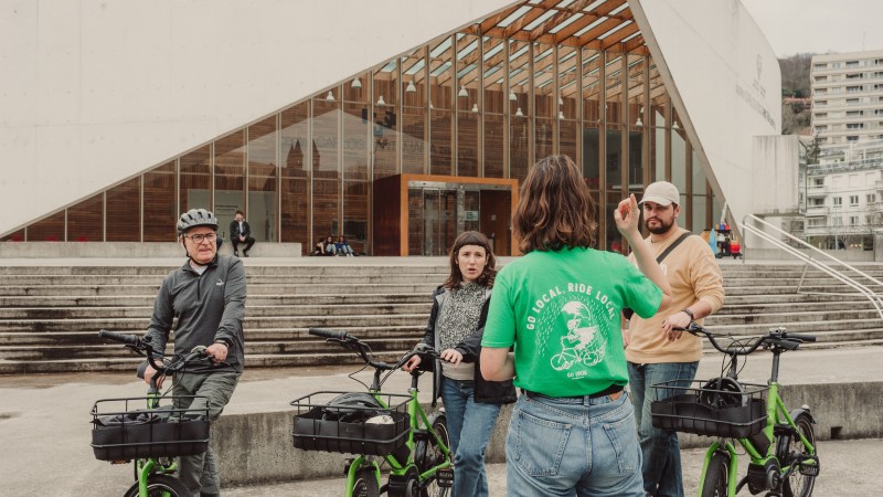 Group of people with bikes in front of a modern building with glass and stairs.